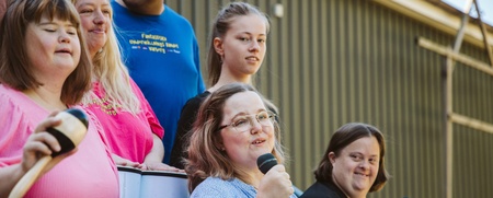 En grupp människor, flera med mikrofon och musikinstrument, står utomhus och sjunger tillsammans.