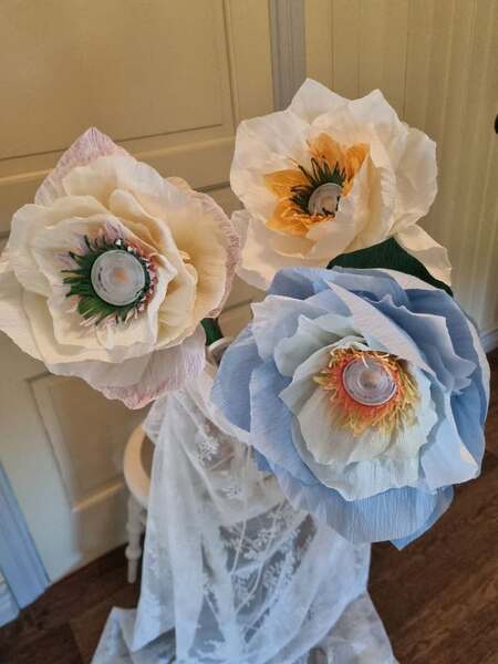 Tre stora konstgjorda blommor av papper med plastlock i mitten, i pastellfärger, står mot en ljus bakgrund.
