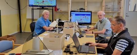 Tre män arbetar vid bärbara datorer i ett kontorsrum med stora bord och en skärm på väggen.