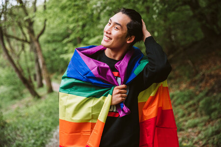 En leende person står utomhus med en regnbågsflagga runt axlarna, omgiven av grönskande träd.