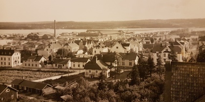 Gammalt svartvitt sepiafärgat foto över en småstad med trähus, fabriker och vatten i bakgrunden.