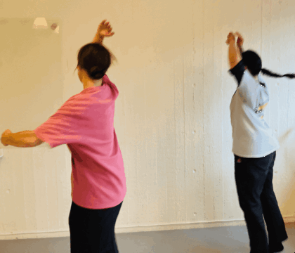 Två personer dansar eller tränar med armarna uppsträckta i ett ljust rum med vit vägg.