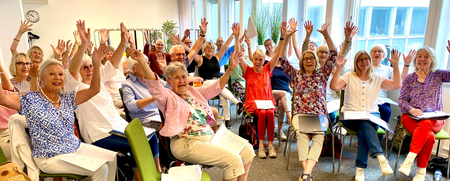 En grupp äldre personer sitter i ett ljust rum och vinkar glatt med händerna i luften.