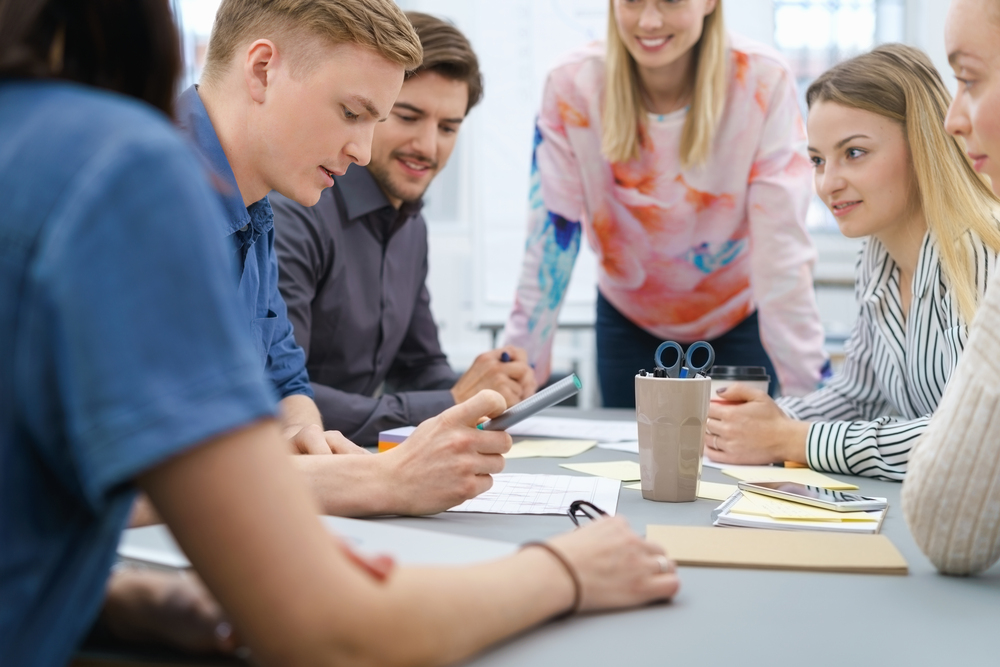 En grupp unga vuxna sitter runt ett bord och diskuterar dokument och en surfplatta.