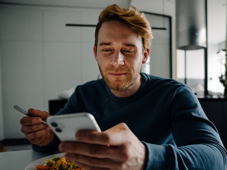 Man sitter vid ett bord, äter mat och tittar på sin mobiltelefon i ett modernt hem.