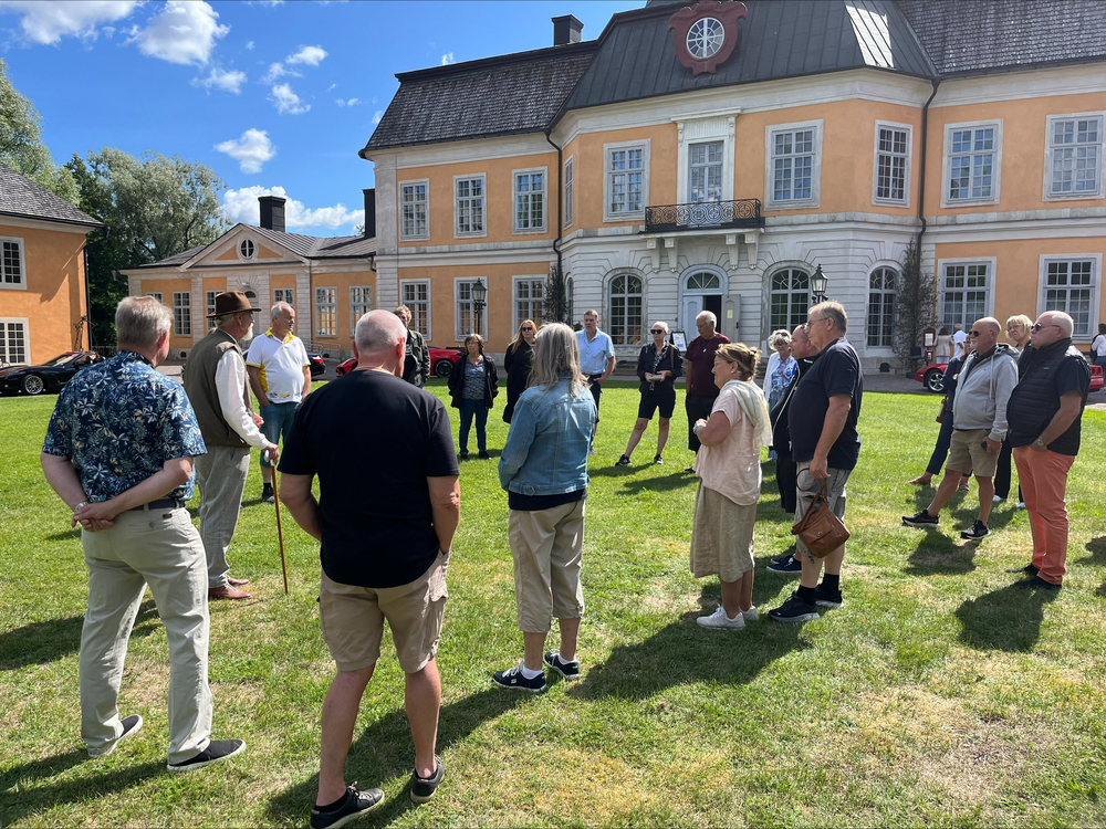 En grupp människor står på en gräsmatta framför en stor gul herrgårdsbyggnad under en solig dag.