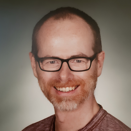 Man med glasögon och skägg ler mot kameran, neutral bakgrund.