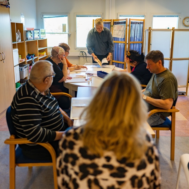 En grupp vuxna sitter runt ett bord i ett klassrum och deltar i en diskussion eller lektion.
