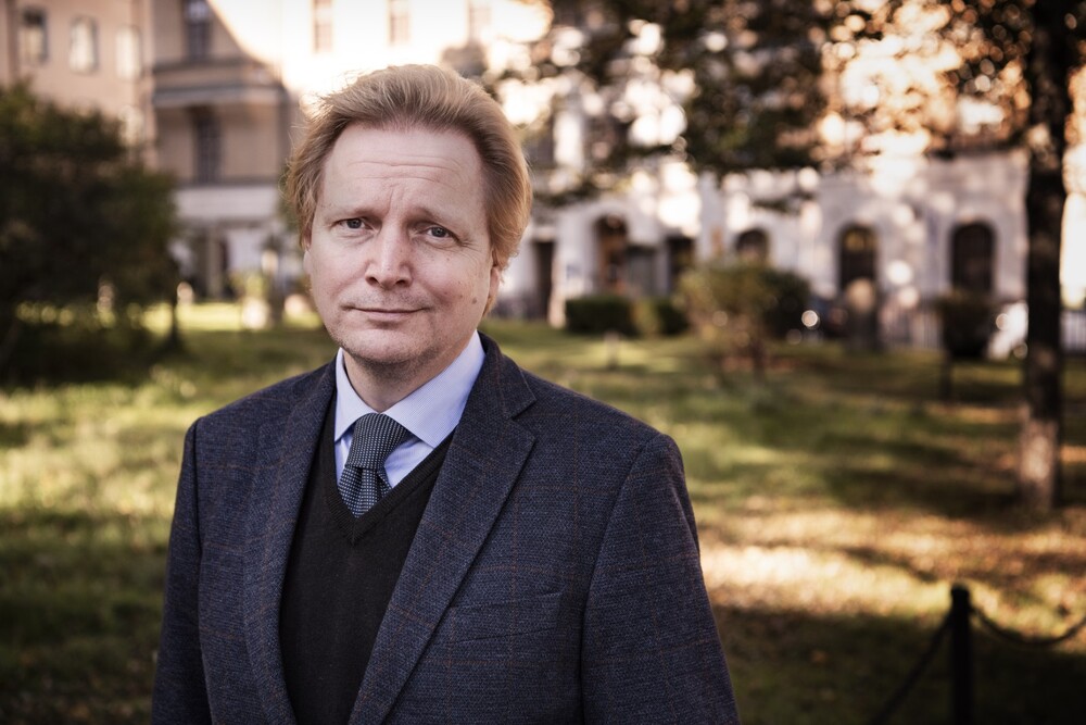 En person i kostym står utomhus i en park med byggnader i bakgrunden en solig dag.