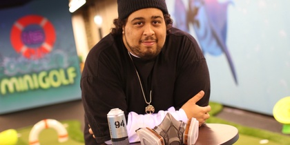 Man med mössa lutar sig mot ett bord med sprayburk och ansiktsmask i en inomhus minigolfmiljö.