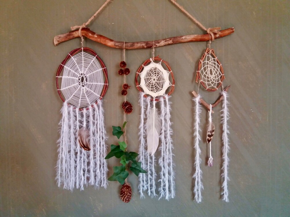 Tre drömfångare hänger på en träpinne mot en grön vägg, dekorerade med fjädrar, garn och kottar.