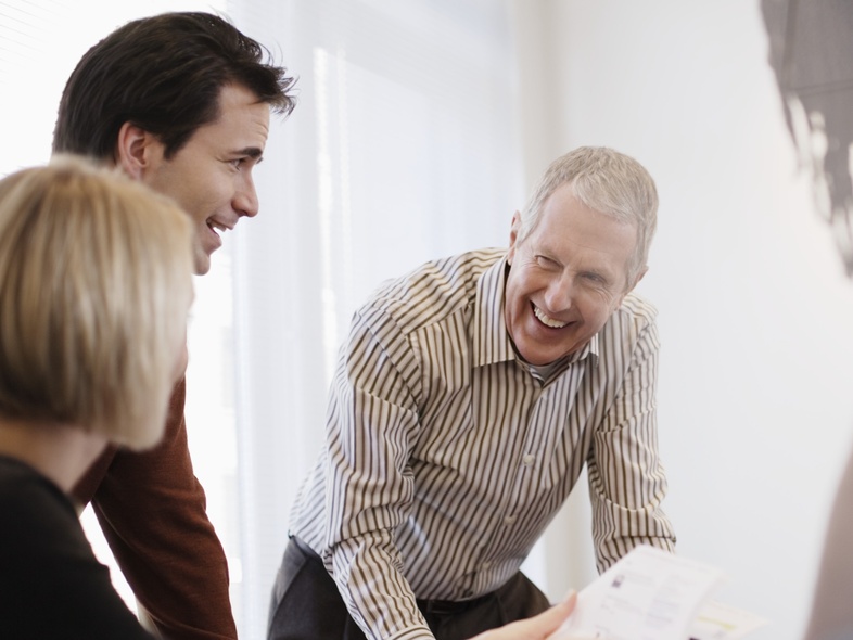 Tre personer har ett glatt samtal runt ett bord i ett ljust kontorsrum.