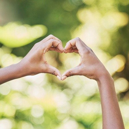 Två händer formar ett hjärta mot en suddig, grön bakgrund utomhus.
