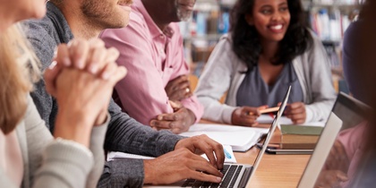 Fyra vuxna personer sitter vid ett bord i ett bibliotek och arbetar tillsammans med datorer och anteckningar.