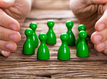 Två händer skyddar en grupp gröna spelpjäser på ett träbord.
