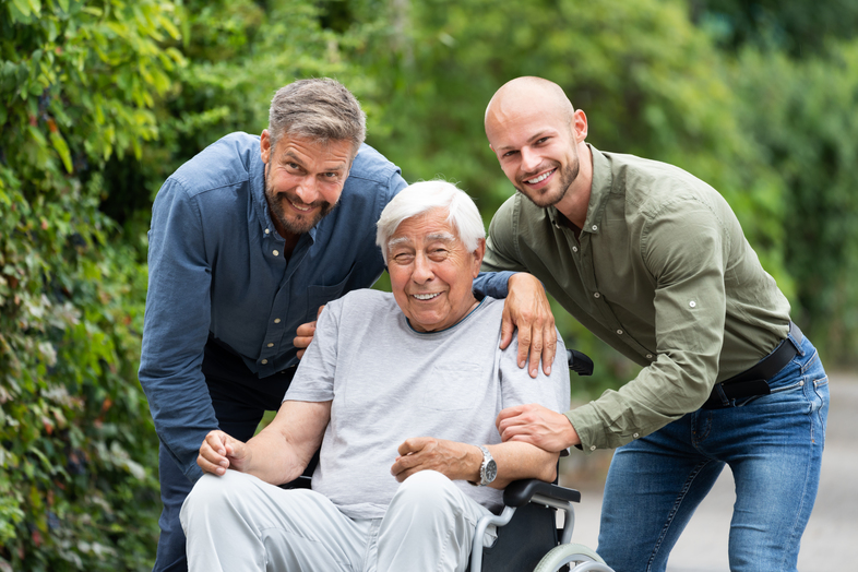 Två yngre män står bredvid en äldre man i rullstol utomhus, alla ler mot kameran.
