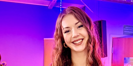 Person med långt lockigt hår håller en bukett blommor i en färgglad inomhusmiljö med lila och rosa belysning.