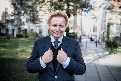 Man i kostym står utomhus på en trottoar med händerna på kavajen och ler mot kameran.