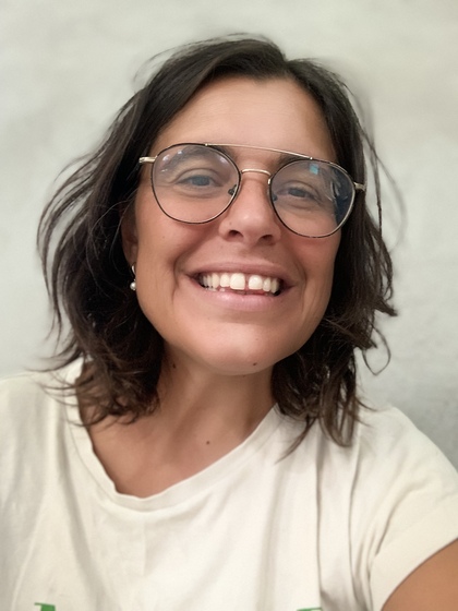 Leende person med glasögon och mörkt, axellångt hår mot ljus bakgrund.