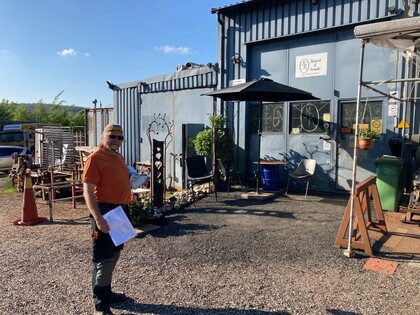 Person med orange tröja står utanför en industribyggnad i solsken, håller papper i handen.