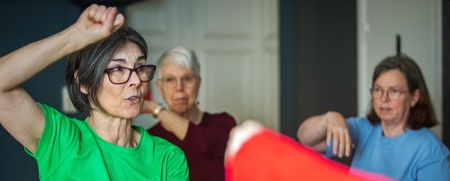 Tre äldre kvinnor deltar i en tränings- eller självförsvarsklass inomhus, en i grön tröja leder övningen.