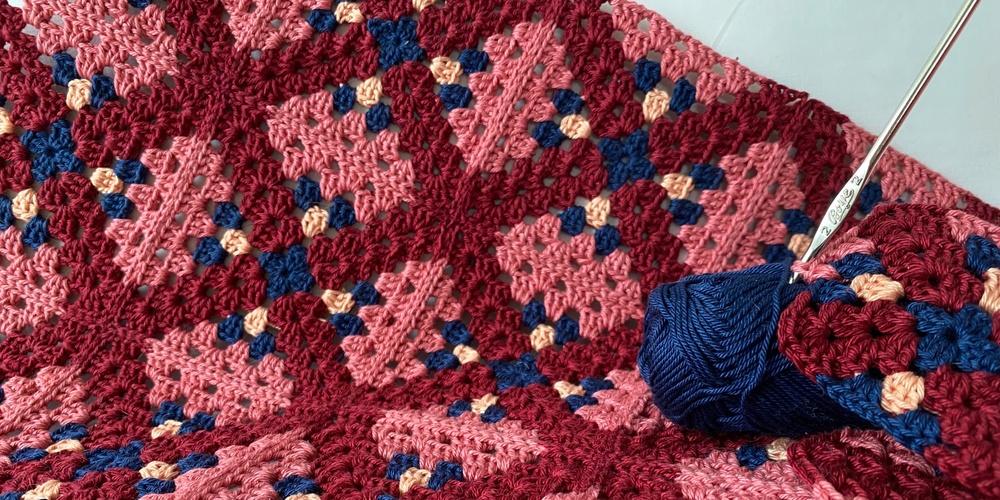 Närbild av ett färgglatt virkat täcke i rutor med garnnystan och virknål.