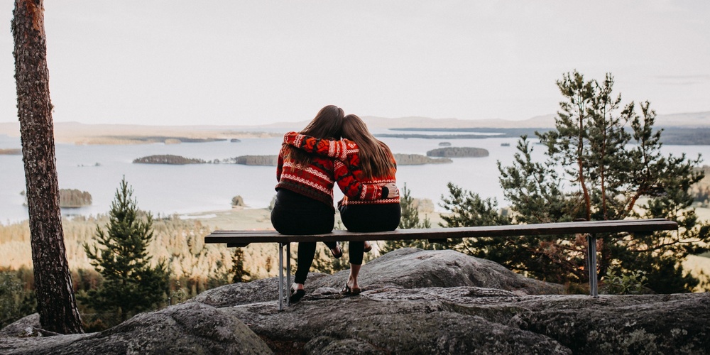 Två personer sitter på en bänk med ryggen mot kameran och ser ut över en sjö och skog.