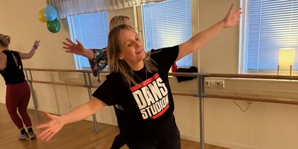 Kvinna dansar glatt i en dansstudio med trägolv och speglar, iförd svart t-shirt med texten 'DANS STUDIO'.
