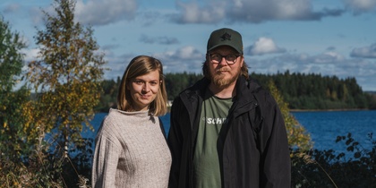 En kvinna och en man står utomhus vid en sjö med skog i bakgrunden under en solig dag.