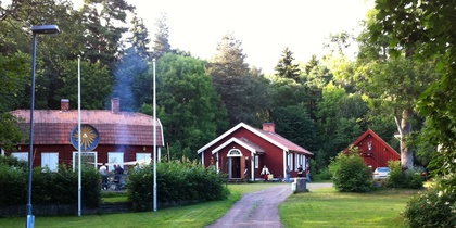 En grusväg leder till tre röda hus omgivna av grönska och träd, med flaggstänger och en grillplats.