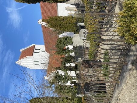 Vit kyrka med rött tak och gravstenar omgiven av järnstaket under blå himmel.