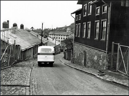 En svartvit bild av en gammal bil som kör nerför en kullerstensgata mellan äldre hus.