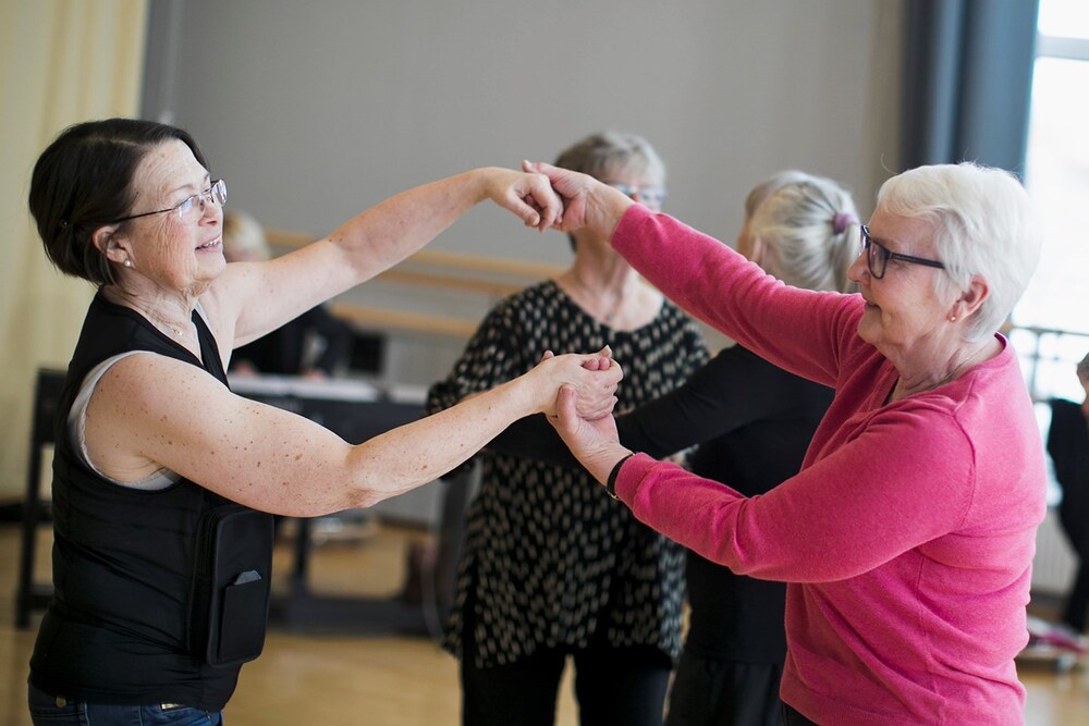Två äldre kvinnor dansar tillsammans i en ljus inomhusmiljö, leende och håller varandras händer.