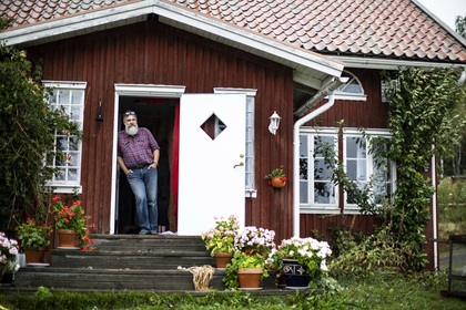 En person står i dörren till ett rött trähus med vita detaljer och blomkrukor på trappan.