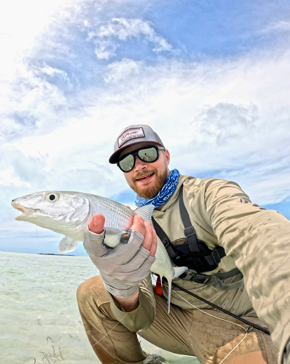 Person i fiskekläder håller en silverfärgad fisk vid grunt vatten under blå himmel.