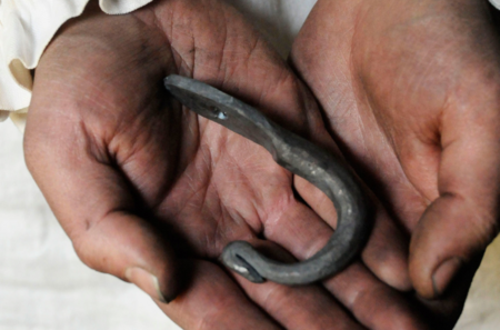 Händer som håller en handsmidd järnkrok, med spår av smuts och arbete på huden.