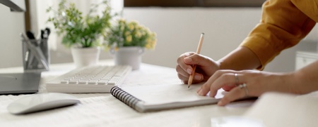 En person skriver i en anteckningsbok vid ett ljust skrivbord med dator, växter och fönster i bakgrunden.