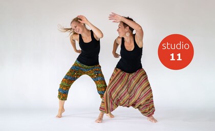 Två kvinnor dansar energiskt i färgglada byxor mot vit bakgrund, med texten 'studio 11' i röd cirkel.
