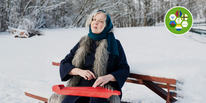 Kvinna i vinterkläder sitter på bänk i snölandskap och spelar ett rött stränginstrument.