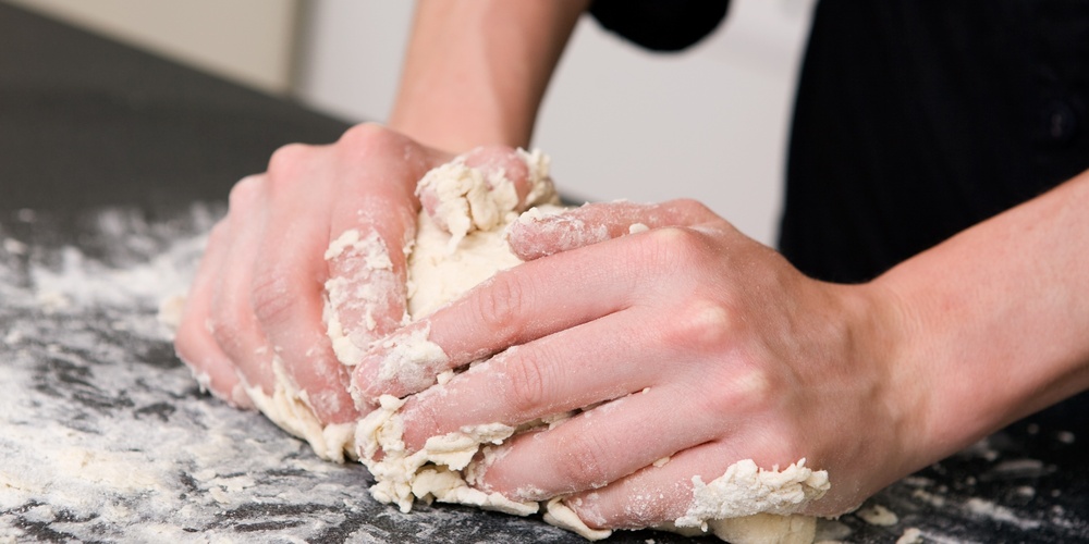 Två händer knådar deg på en mjölad bänk i ett kök.