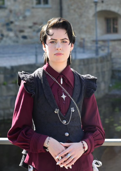 En person med kort mörkt hår och smycken bär en vinröd skjorta och svart väst, står utomhus framför en stenbyggnad.