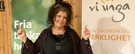 Kvinna står leende framför två rollups i ett rum med träpanel och pekar uppåt med båda händerna.