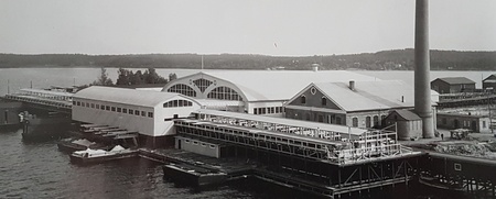 Svartvit bild av en industribyggnad vid vattnet med stor skorsten och bryggor.