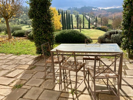 Ett glasbord med fyra stolar står på en stenlagd uteplats med utsikt över ett grönt landskap och cypresser.