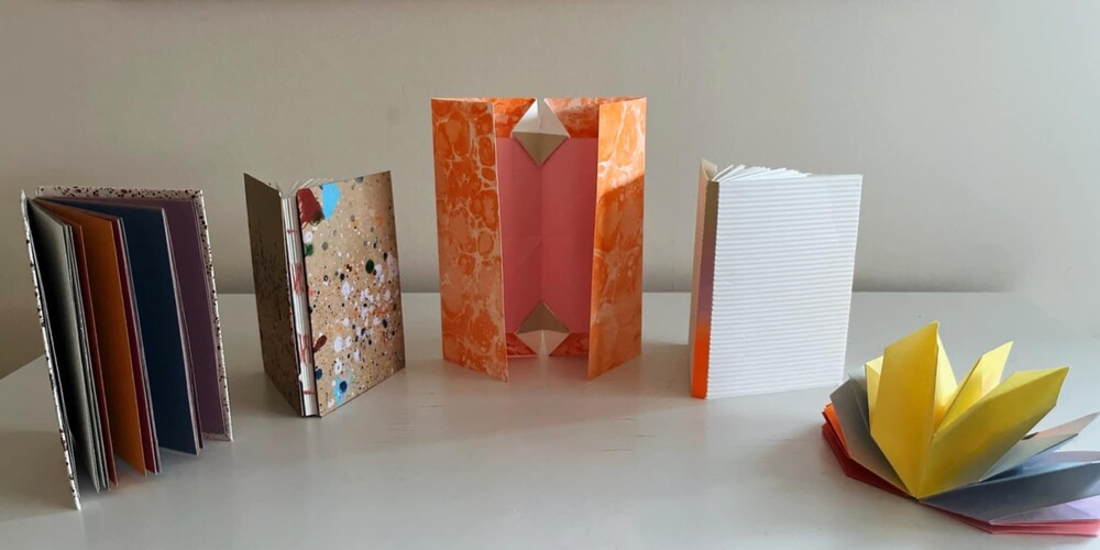 Fem färgglada handgjorda böcker och vikta papper står på ett vitt bord mot en ljus vägg.