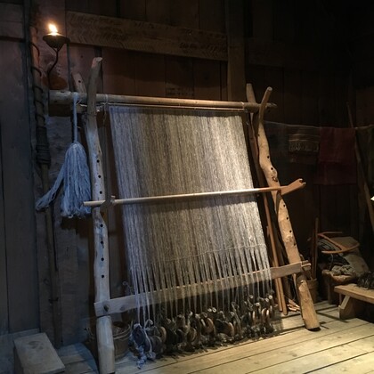 Ett gammaldags vävstol i trä står i ett dunkelt rum med trägolv och väggar.