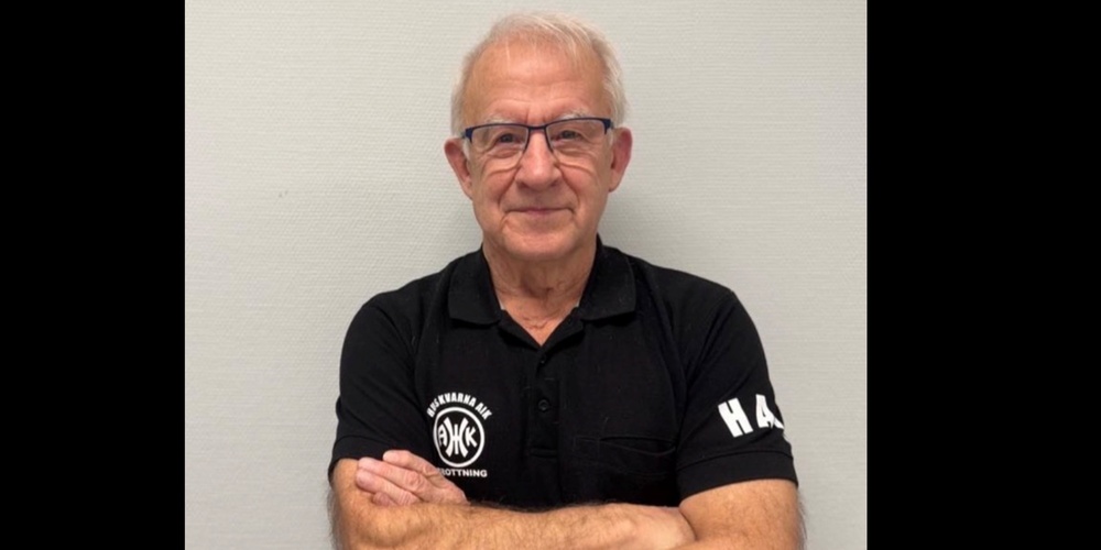 Man med armarna i kors, iförd svart pikétröja med vit text, står mot ljus vägg. Ansiktet är suddat.