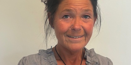 Kvinna med mörkt hår i uppsättning, grå blus med volang, sitter på svart stol mot ljus bakgrund.