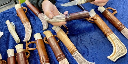 En person visar handgjorda knivar med dekorerade slidor på ett blått tygbord.