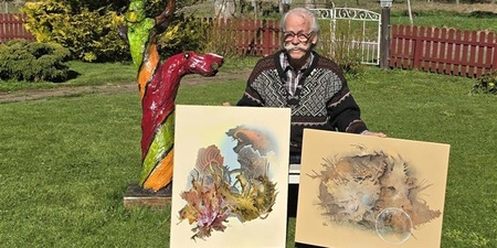 Person sitter utomhus och visar två konstverk, med en färgglad träskulptur bredvid.